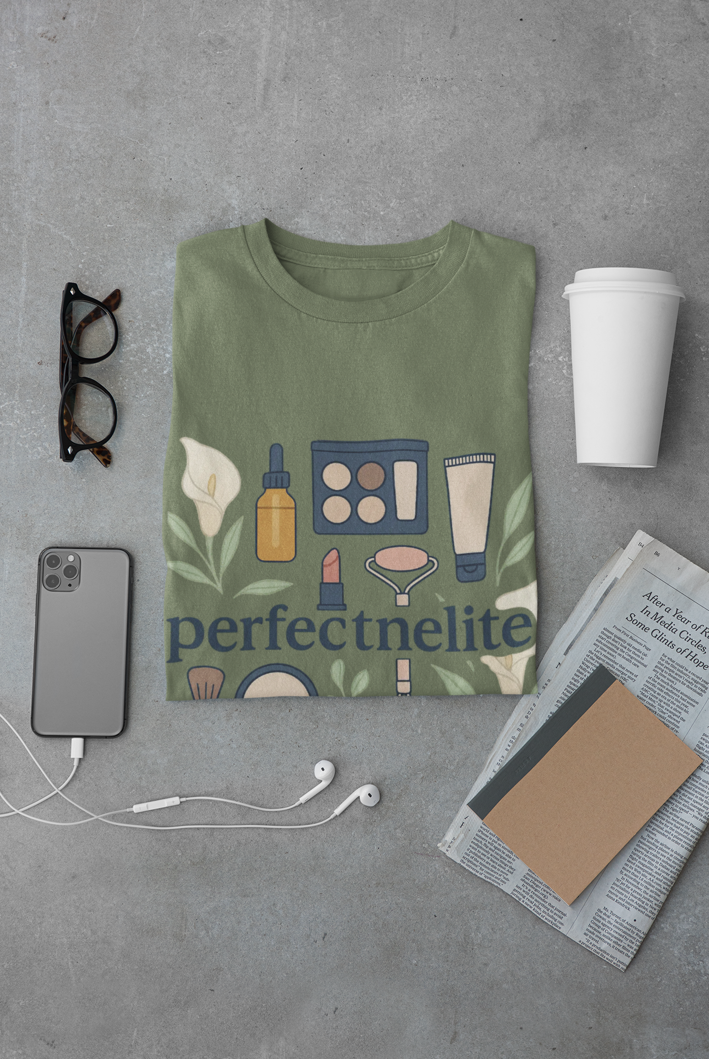 Folded green Perfectnelite graphic t-shirt with beauty and skincare icons, styled flat lay with coffee cup, phone, glasses, earphones, and newspaper.